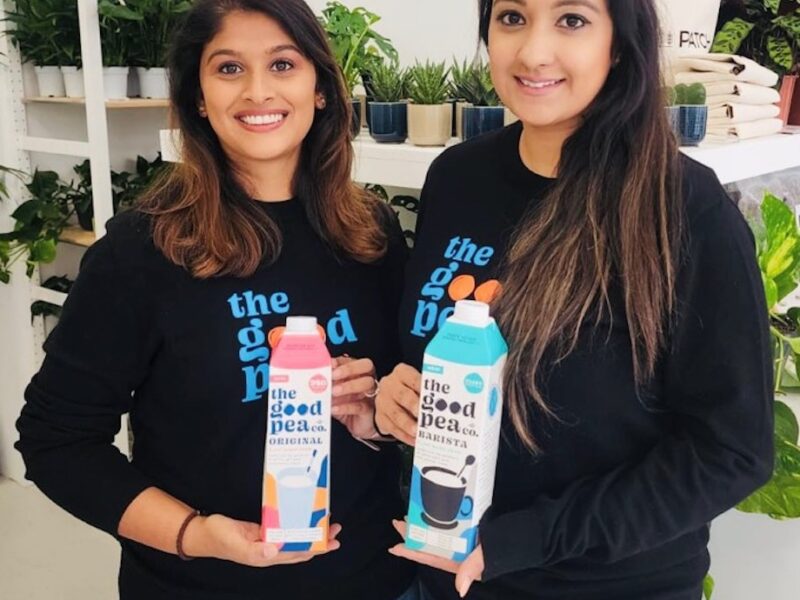 Two young women holding 'The Good Pea Co' milk cartons and smiling, with potted plants in the background