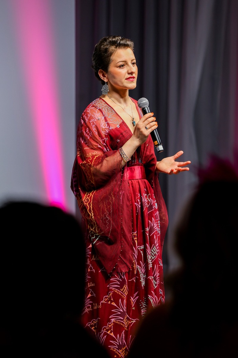 A woman with short hair in a red dress, onstage and speaking into a microphone