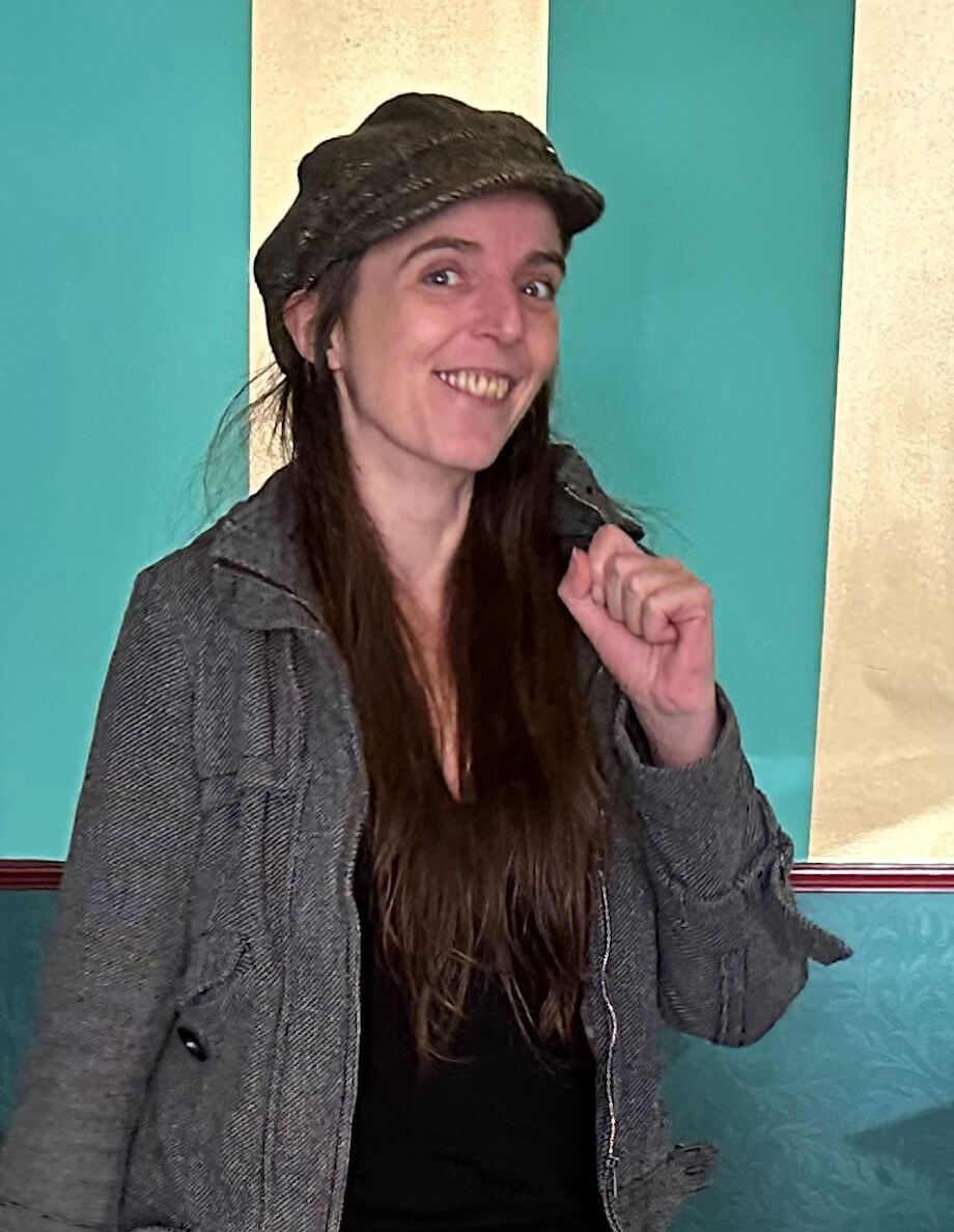 A woman smiling, wearing a hat and coat, against a turquoise background