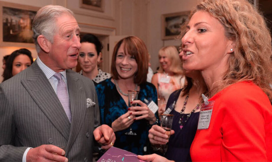 A woman in a bright red shirt holding a book and talking to King Charles, guests standing in the background, smiling with drinks